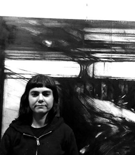 A black and white portrait of Raha Raissnia in front of an abstractly drawn black and white background. She has short black hair and bangs and wears a black zip-up sweatshirt. 