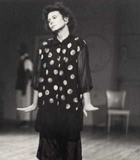 A black and white portrait of Susan Rethorst wearing a polka dot shirt and a black skirt with her hair in an up do. She looks down and to the left and holds her arms beside her body as her hands flare out to the sides. The background of the room behind her is blurry but a metal chair and wooden floor is visible.