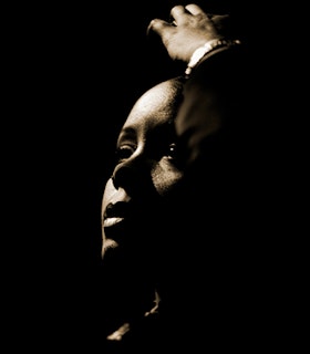 A dark lit side angle portrait of Matana Roberts with their hand above their head and their profile accented by shadows. 
