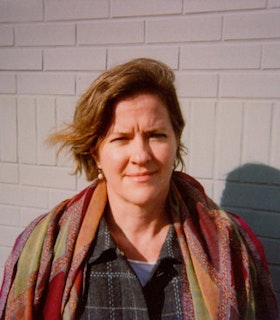A portrait of Elizabeth Robinson in front of a grey brick wall. She has short brown hair which is swept to the side and wears a multicolored scarf. 