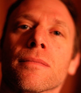 A close up, red tinted portrait of Steve Roden. He has short hair and stubble on his chin and upper lip. 