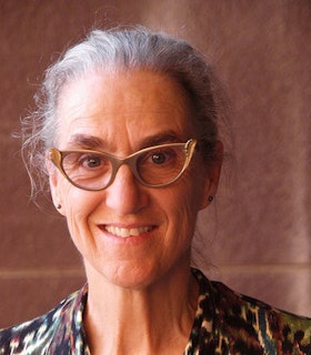 A close up portrait of Wendy Rogers smiling in front of a blurred burgundy background. She wears tan cat eye glasses and a green and purple patterned blouse. Her grey hair is pulled into an updo. 
