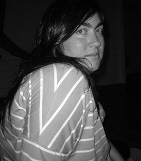 A black and white portrait of Jen Rosenblit against a black background. She has medium length black hair and wears a white and grey striped shirt. While her body angled to the side, she faces the camera directly. 