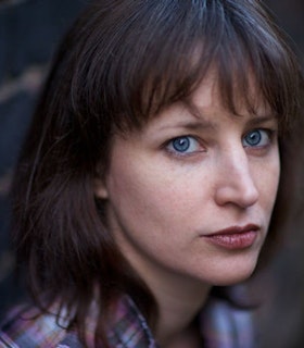 A portrait of Marina Rosenfeld in front of a blurred grey background. She has medium length brown hair and bangs and wears a blue and pink flannel shirt.
