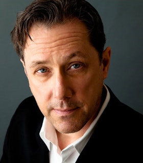 A close of portrait of Mikel Rouse in front of a dark grey background. He wears a white shirt and black suit jacket and has short brown hair. He tilts his head slightly to the left and raises his right eyebrow, smiling slightly.