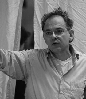 A black and white portrait of Sinan Savaskan in front of a hung wrinkled white sheet. He wears a button down shirt over a white tank top and thin wire rimmed glasses. Lifting his right arm up and looking towards it with his mouth somewhat agape, he appears to be directing someone. 