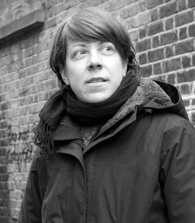 A black and white portrait of Grace Schwindt in front of a brick wall. She has short hair and wears a dark parka and scarf. 