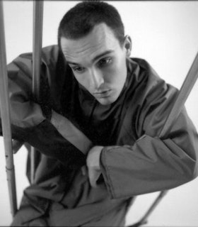 A close up black and white portrait of Bill Shannon supporting himself by the arms on metal crutches. He wears a grey tunic and has a closely shaven head. He tilts his head downwards and appears lost in thought.  