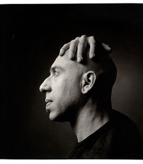 A black and white portrait of Elliot Sharp. He is bald and wears a black shirt. His profile faces the camera and as he looks forward, he reaches around to hold the top of his head with one hand.