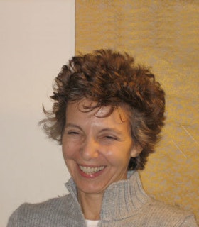 A portrait of Vicky Shick smiling towards her lower right side against a half wooden half beige background. She wears a grey turtleneck jacket and has short curly hair.