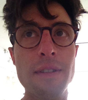 A close up image of Gedi Sibony against an overexposed bright background. He has short brown hair and wears circular tortoise shell glasses. He looks upwards and opens his mouth slightly. 