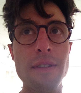 A close up image of Gedi Sibony against an overexposed bright background. He has short brown hair and wears circular tortoise shell glasses. He looks upwards and opens his mouth slightly.