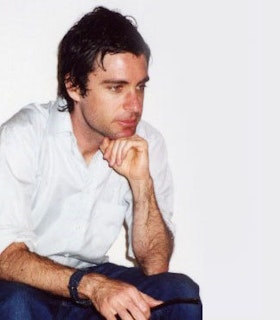 A profile portrait of Paul Sietsema in front of a white background. He wears blue pants and a white shirt. Sitting down and resting his chin on his hand, he looks out a something beyond the frame of the image with a thoughtful expression on his face. 