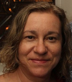 A close up portrait of Sally Silvers smiling and looking directly at the camera. She has shoulder length flax colored wavy hair. Behind her, there is a window which overlooks a dark scene with numerous lights within it. 