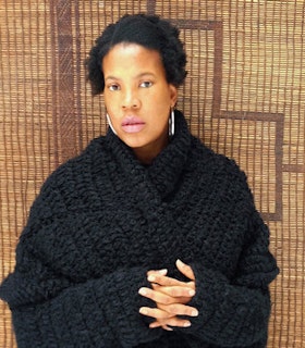 Portrait of Xaviera Simmons with short dark hair, hoop earrings, and a chunky black sweater. Their fingers are interwoven.
