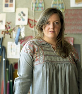 A portrait of Allison Smith in front of a blurred wall with drawn pictures hung on it. She has long brown hair, wears a grey and red embroidered shirt, and is looking directly at the camera. 