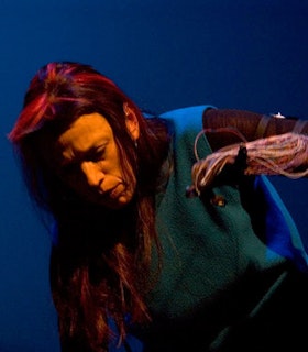 Laetitia Sonami leans over in front of a blue background, appearing to be mid-performance. She wears a blue fleece vest and has long hair with bright fushia streaks in it. She closes her eyes slightly and looks downward with a concentrated expression on her face. She lifts her left arm up to her face. That arm is covered in numerous thin wires.