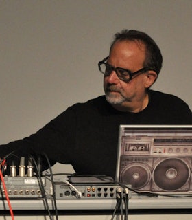 A portrait of Carl Stone working at a sound machine against a gray background. He has a beard and short hair and wears a dark shirt and black glasses. He looks towards a sound machine on his left. 