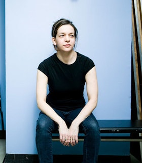 A portrait of Pam Tanowitz sitting on a black bench in front of a light blue background. She wears blue jeans and a black t-shirt and has her hair pulled into a low ponytail. Her elbows rest on her thighs and she looks beyond the camera towards the upper right. 