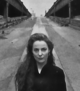 A black and white portrait of Fiona Templeton standing in front of a long road. She has long hair and wears a dark top. She looks directly at the camera with an intent expression on her face.   