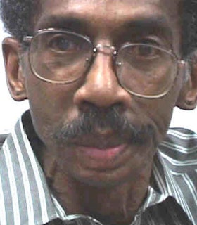 A close up portrait of Lorenzo Thomas in front of a white background. He wears a blue and white striped shirt and tilted wire glasses. He has short hair and a mustache. He looks directly at the camera and doesn't smile. 