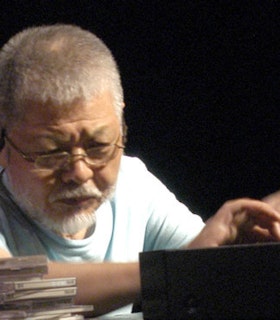 A close-up portrait of Yasunao Tone. He has short grey hair and grey facial hair, think wire glasses and wears a light blue t-shirt. Beside him, there is a stack of CD cases. His hands gesture a top a dark box in front of him.