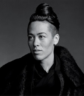 A black and white side angle portrait of Wu Tsang against a grey background. She has an undercut and a high bun and is wearing a black fur jacket. 