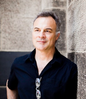 Portrait of Toby Twining against a stone tiled corner. He has short grey hair and wears a navy blue short sleeve collared shirt with a pair of glasses hanging from his neckline. 