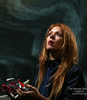 A portrait of Kate Valk in front of a turquoise background with a pattern of wavy lines on it. She has long red hair and wears a dark blue top. She looks up with an expression of awe on her face and holds a contraption made of red and silver wires and trinkets.