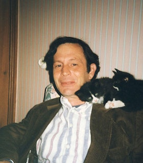 A close up portrait of Paul Violi in front of a pink and white striped wall. He wears a purple and blue striped shirt and a brown suede jacket and looks directly at the camera. Two black and white kittens sit on his shoulder.