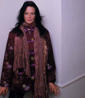Anne Waldman stands in front of the corner of a room looking intently at the camera. She has chest length dark hair and wears a floral patterned purple and green dress and a long salmon colored scarf. 