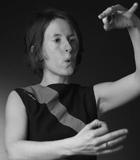 A black and white portrait of Jennifer Walshe, framing her face with her arms and holding her hands in front of her, with her pointer finger and thumbs held together, making a circle. She puckers her lips, appearing mid-whistle. 