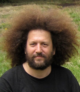 A portrait of Michael Webster in front of a grassy field. He has medium length light brown hair, a thick dark brown beard, and wears a black shirt. 