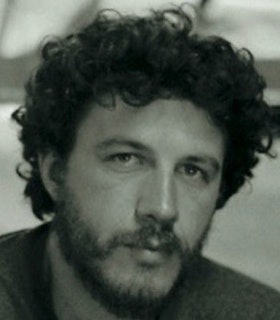 A black and white portrait of Ishmael Randall Weeks in front of a blurred background. He has dark curly hair and stubble beard.