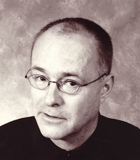 A black and white, pink-hued portrait of Mac Wellman wearing a black top and thin, wire-rimmed glasses. He looks directly at the camera despite turning his head in a three quarter profile. The background of the image is a blurred pattern of a dark and light shade blended together.