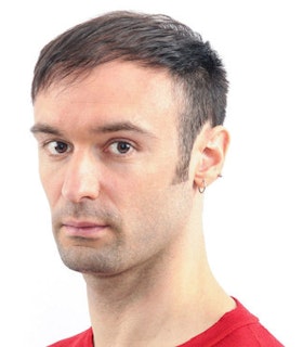 A 45-degree angle portrait of Christopher Williams looking into the camera against a white background. He wears a red shirt and has dark short hair.