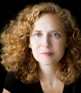 A close up portrait of Julia Wolfe in front of a dark background. She wears a dark shirt and has curly red hair. She looks directly at the camera and doesn't smile. 