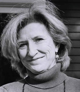 A close up black and white portrait of C.D. Wright wearing a turtle neck and a beaded necklace. She has shoulder length hair and smiles as she looks at the camera.