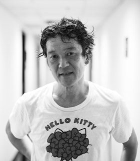 A black and white portrait of Kota Yamazaki in front of a blurred white background. He has short curly black hair and wears a hello kitty t-shirt.