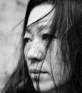 A black and white, side angled portrait of Yasuko Yokoshi looking into the distance. She has black hair which is blown into her face. 