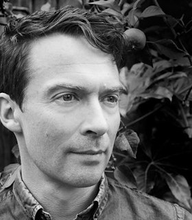 A black and white close-up portrait of Adam Chodzko in front of a leafy tree. He is in three quarter profile and looks out at someone beyond the frame of the image. He has short dark hair and wears a collared shirt. 