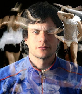 A portrait of Foofwa d'Imobilité in front of a black background. He has short black hair, blue eyes, and wears a blue zip-up shirt with red accents. Blurred, translucent images of dancers in various positions overlay his portrait at various angles.  