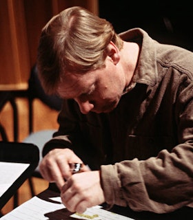 A side portrait of Michael Pisaro dressed in a khaki color jacket. He looks downs and studies a silver object placed on a flat surface.