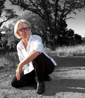 A portrait of Ann Carlson wearing a white button down shirt, black pants, black shoes, and blue glasses. She crouches on a grassy floor and looks to her right at something beyond the camera. Only her body is in color but the background of grass and trees is all black and white. 
