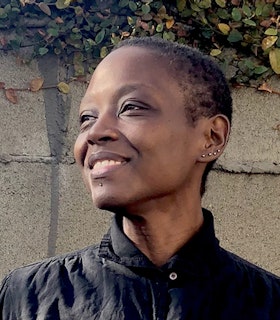 Portrait of taisha paggett with short hair and multiple earings on the ear and one between the chin and mouth, dressed in a black button up. The artist stands, their body en face but their head turned to the side smiling, in front of a wire fence surrounding a wall with greenery on the top.