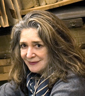 A close up portrait of Rochelle Feinstein. She has shoulder length brown hair and wears a gray top and a blue and navy striped scarf. She looks towards the camera and smiles. 