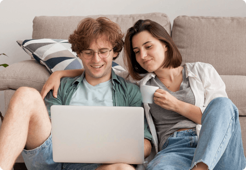 A young couple sitting comfortably on a couch. The man has a laptop on his lap. He is smiling as he looks at the screen. The woman sitting next to him has her arm around his shoulder and is holding a white mug in her other hand.