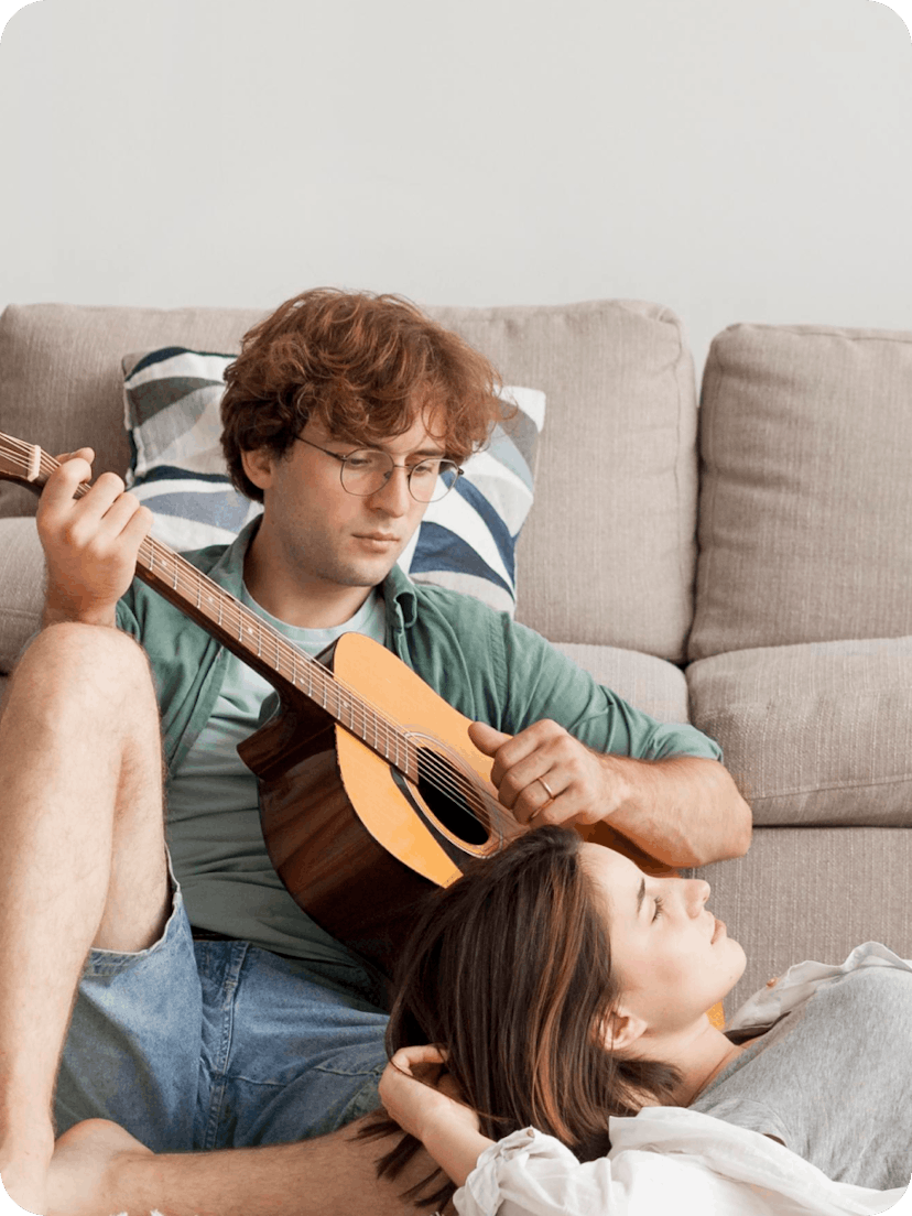 A young couple relaxing together. The man is playing an acoustic guitar while the woman rests her head on his leg, lying down with her eyes closed.