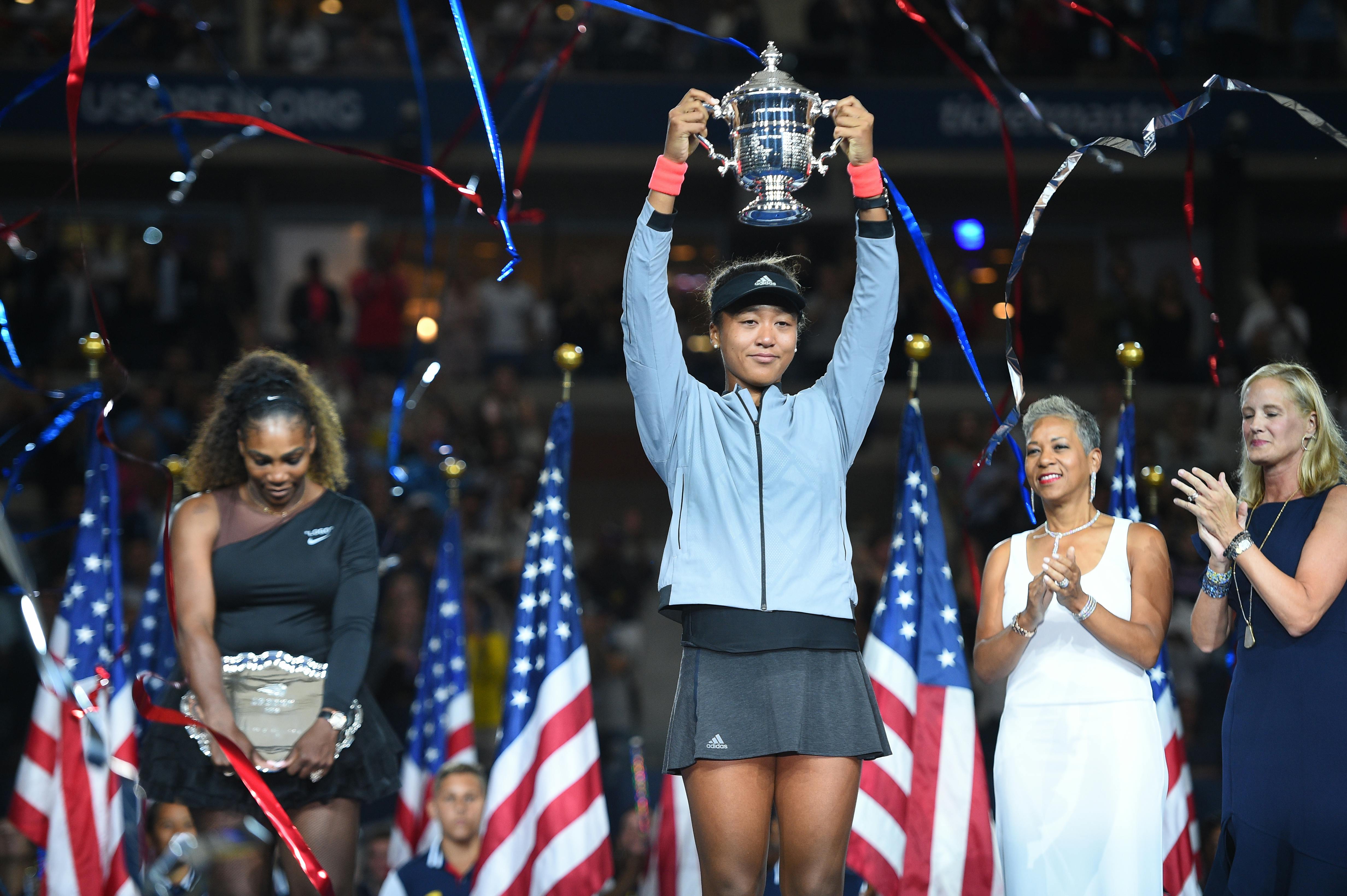 Naomi Osaka holdinh her trophy with Serena Williams in the background US Open 2018