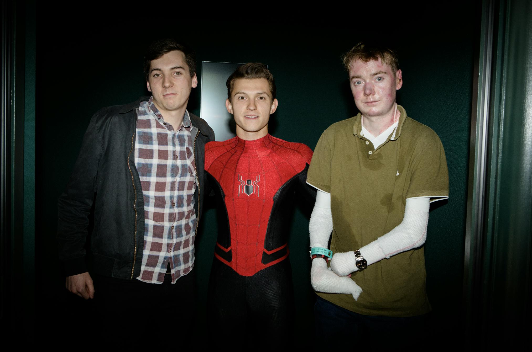 Actor Tom Holland dressed as Spiderman standing between two men - one in a check shirt and one in an olive shirt.